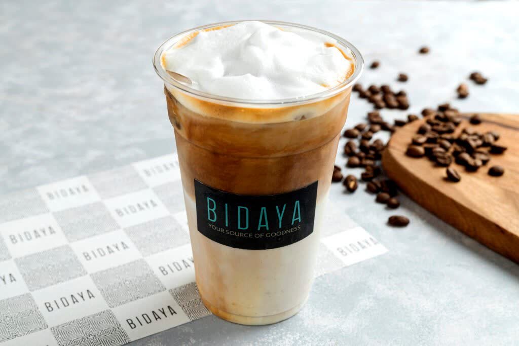 Iced latte with a thick foam cap in a Bidaya branded plastic cup, coffee beans beside