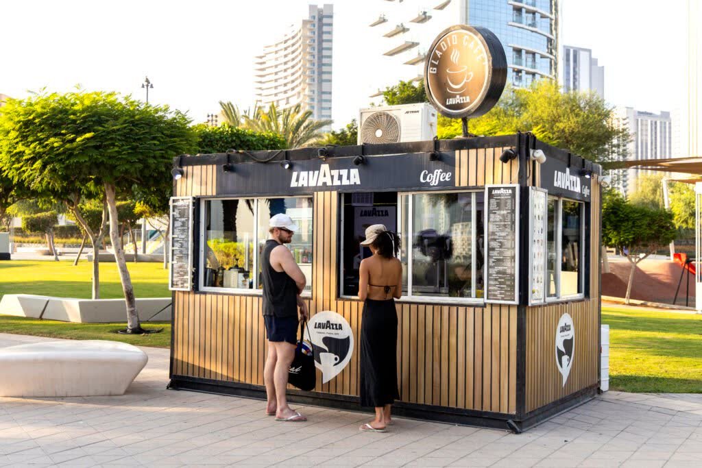 Customers ordering at a branded outdoor coffee kiosk in Dubai, with logo signage above the counter