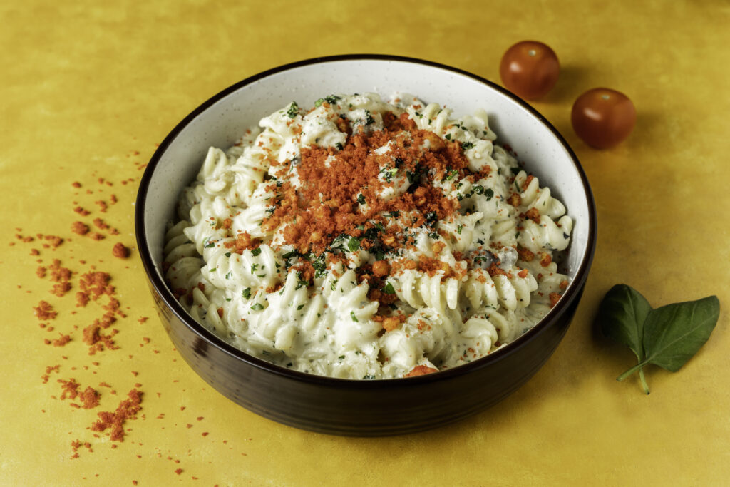 Creamy fusilli pasta in a white sauce topped with red seasoning and fresh herbs, styled for food photography on a yellow background