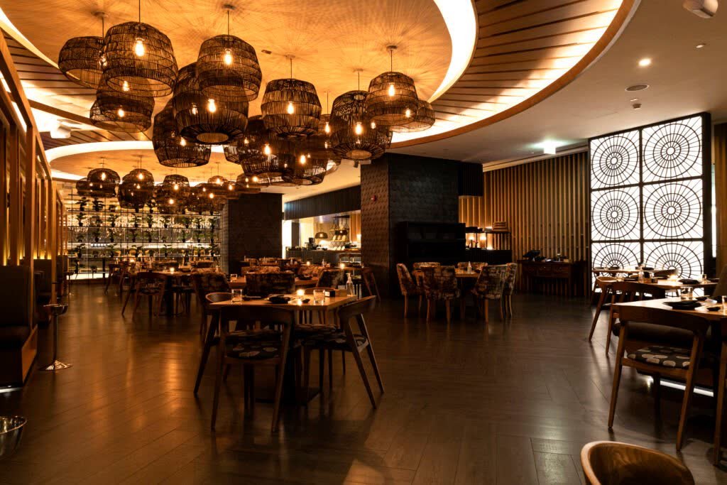 Wide view of a hotel restaurant dining room with woven pendant lights, laid tables and an open kitchen behind