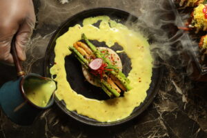 Gourmet plated dish with asparagus and vegetables on a black plate, surrounded by a creamy yellow sauce being poured, with steam rising and a hand holding a sauce pitcher.