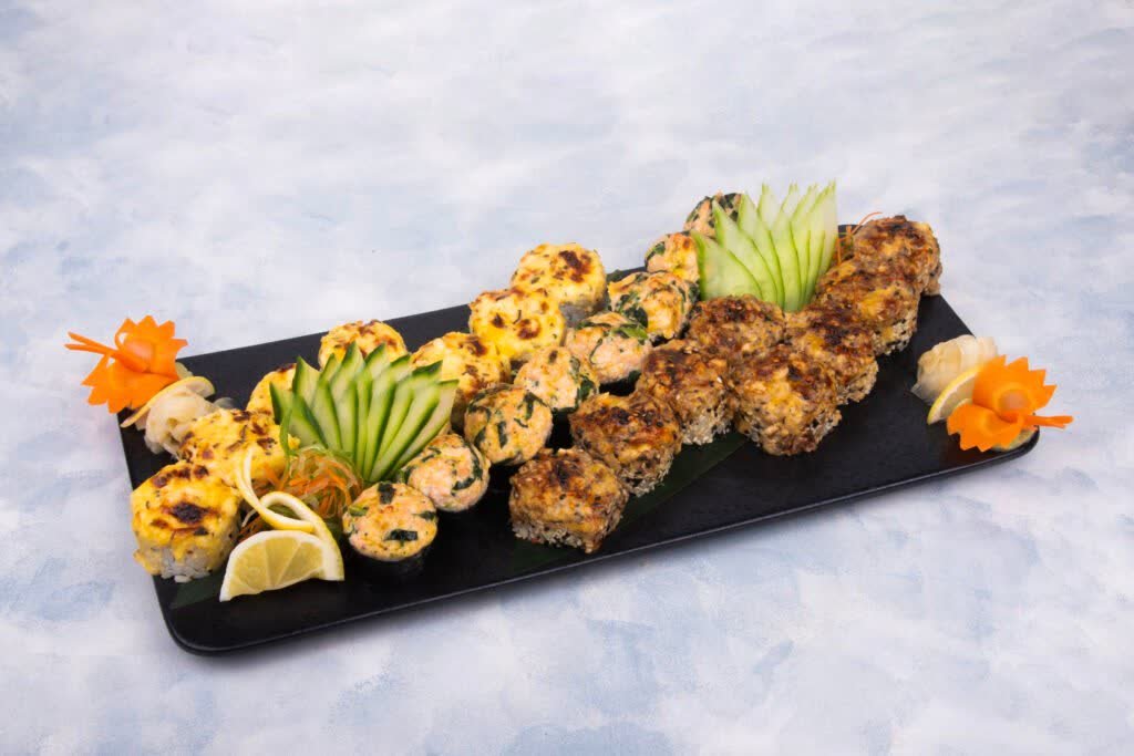 Sushi rolls with browned toppings on a long black platter, garnished with cucumber and carrot