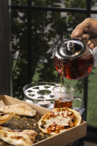 Restaurant menu photography showing hot tea being poured into a glass cup alongside freshly baked flatbreads and mini pizzas, styled in natural light with a cozy dining setting.