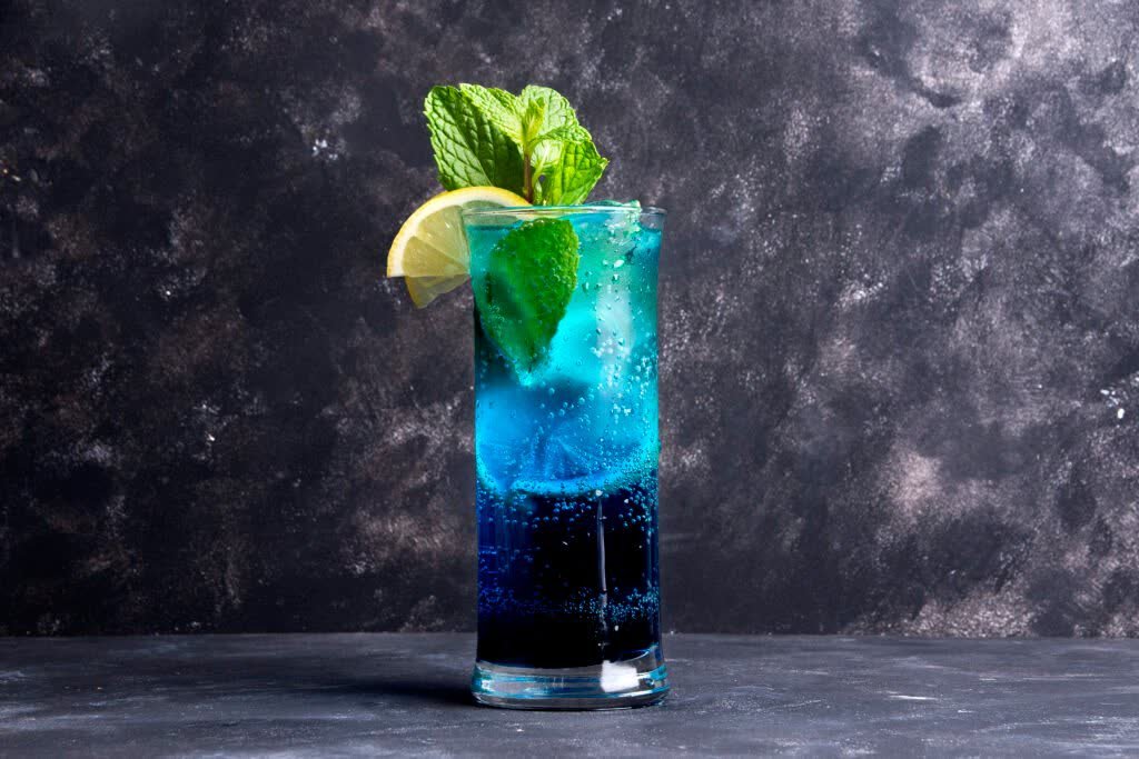 Layered blue sparkling drink with ice, mint and a lemon wedge on a dark backdrop