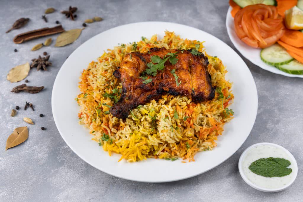 Chicken biryani topped with a roasted chicken leg, beside raita, salad and whole spices