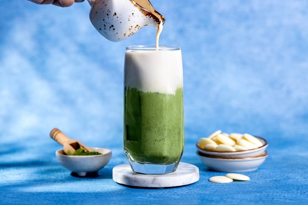 Hand pouring white chocolate foam from a speckled jug into a tall glass of matcha