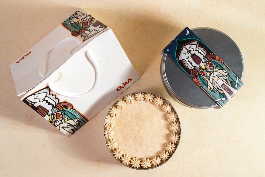 OMA illustrated gift box and dessert tin photographed overhead beside a cream-topped cake