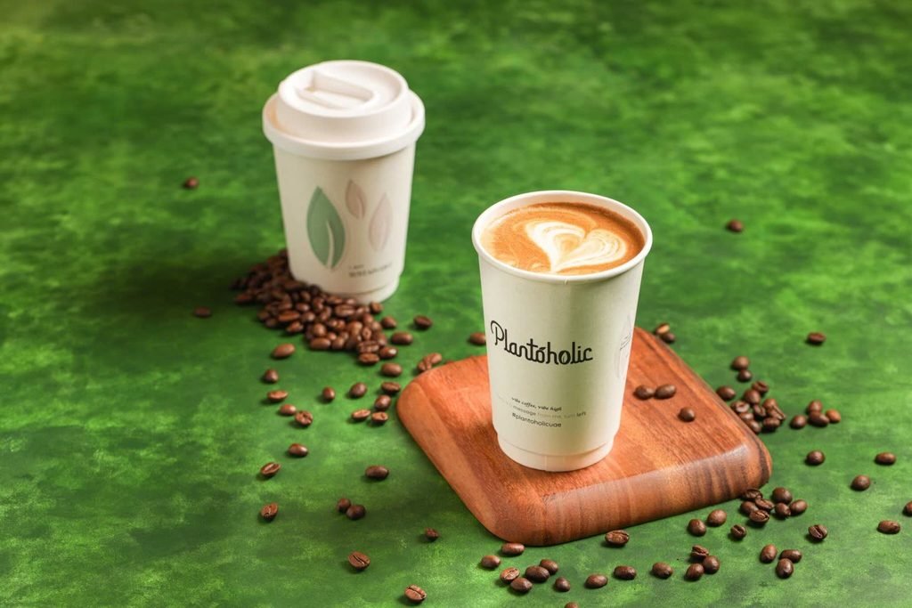 Plantoholic paper cup with heart latte art beside a lidded takeaway cup and coffee beans