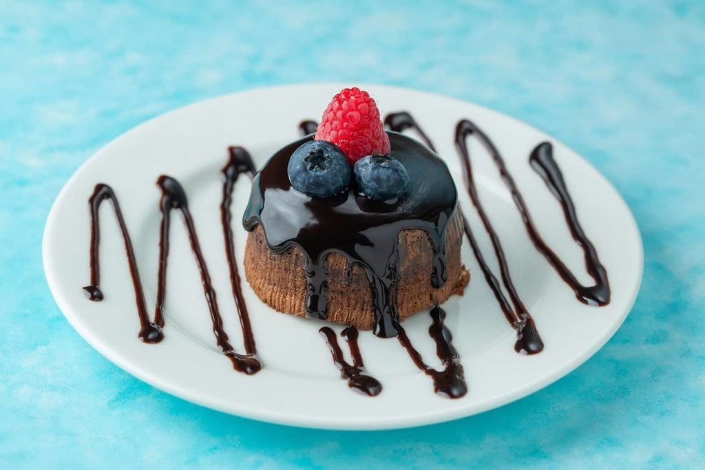 Small chocolate cake glazed with ganache, topped with a raspberry and blueberries, on a white plate