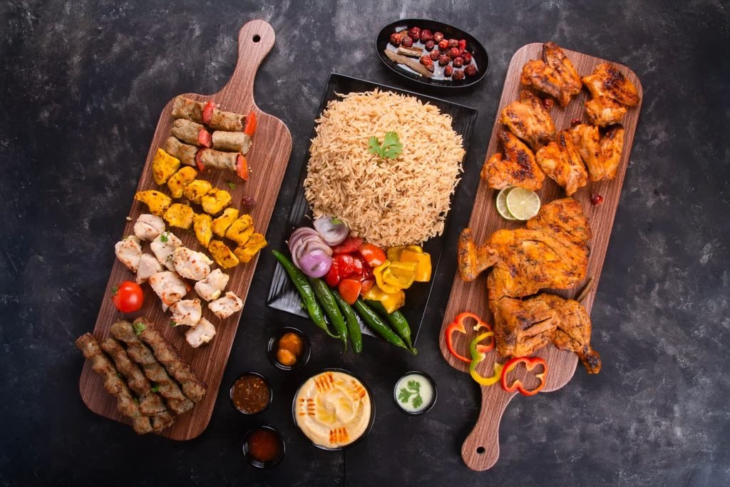 Overhead spread of kebab and grilled chicken platters, rice and dips on a dark table